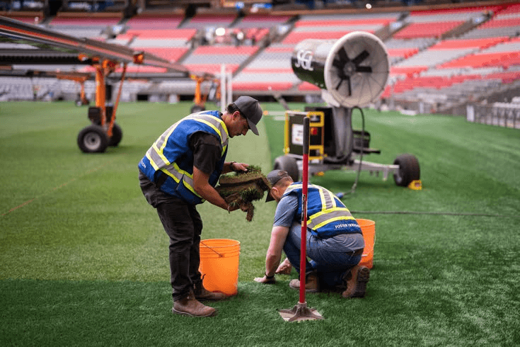 Stadium grounds crew maintaining turf on a sports field with seating in the background
